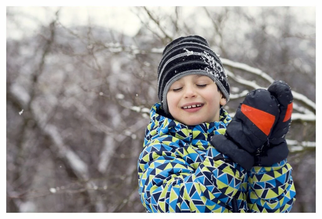 Young boy standing in front of winter trees. There is snow on his wooly hat from a recent snowball. His hands are protected from chapping with thick, waterproof mittens.