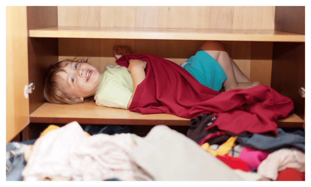 Cheeky child hiding on a shelf in their clothes cupboard. Their clothes are on the floor in the foreground.