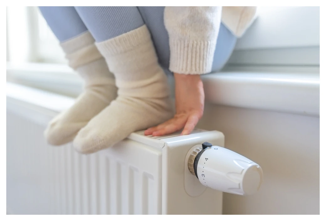 Young child sitting on a windowsill with her feet (in wooly socks) and hands resting on the radiator below. She has just turned up the thermostat.