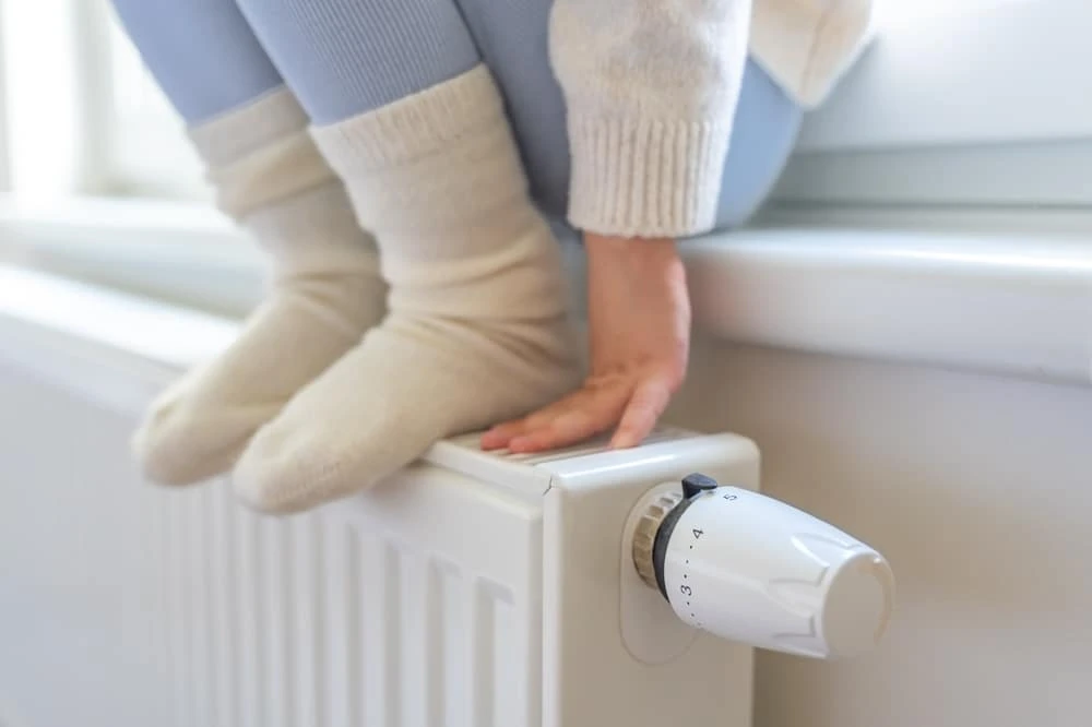 Young child sitting on a windowsill with her feet (in wooly socks) and hands resting on the radiator below. She has just turned up the thermostat.